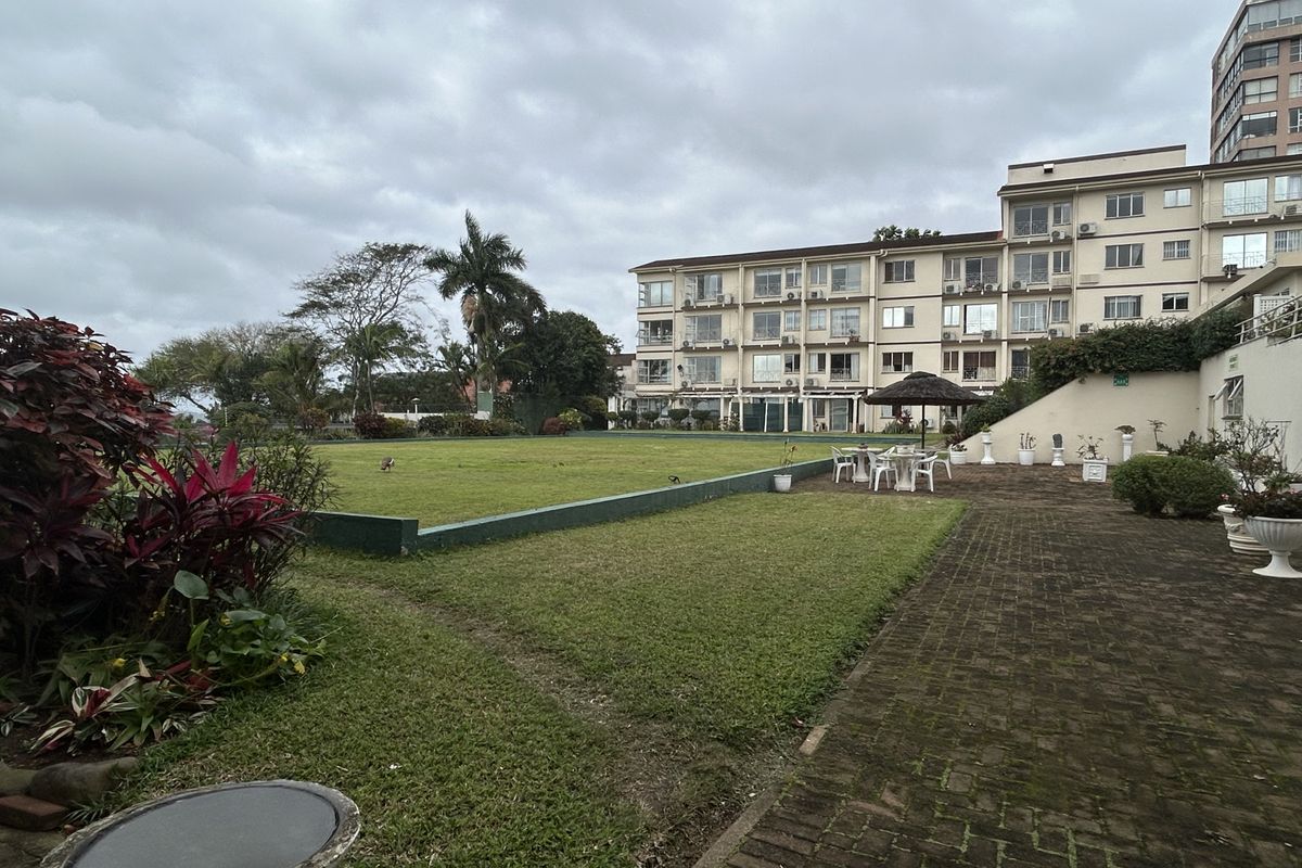 A view of the rear exterior of the retirement village, featuring a paved courtyard area, a large landscaped lawn, and the multi-story residential building.