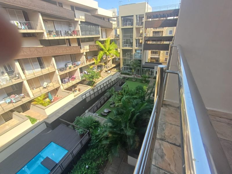 Elevated view from a balcony overlooking the apartment complex's communal garden, swimming pool, and surrounding residential architecture.