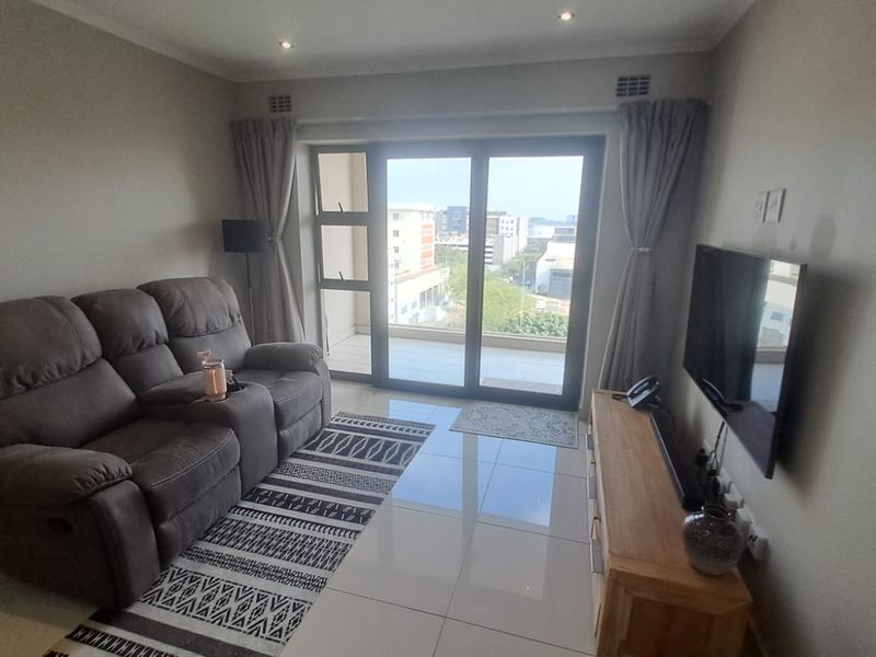 A well-maintained living room featuring polished tile flooring, recessed lighting, and large sliding glass doors providing access to a balcony with urban views.