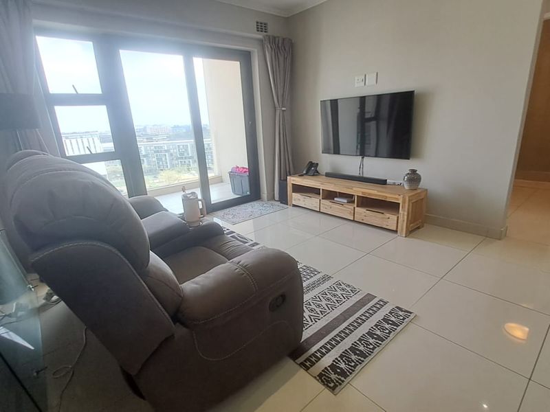 A bright living room featuring large sliding glass doors leading to a balcony, neutral tiled flooring, and recessed ceiling lighting.