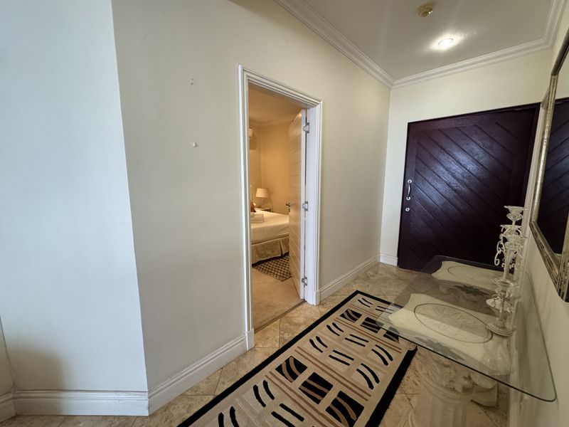A well-lit hallway featuring tiled flooring, crown molding, and a solid wood door leading into a bedroom.