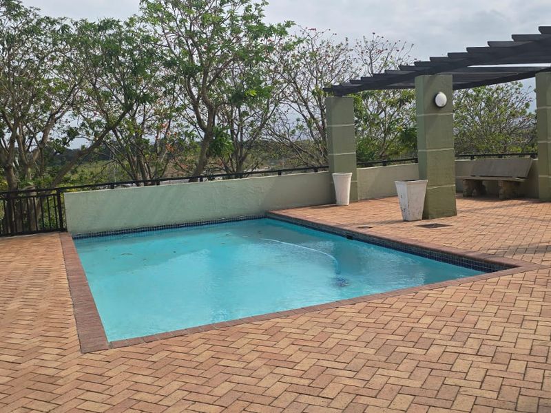 Outdoor pool area featuring a swimming pool, brick paved deck, and a pergola structure, set against a backdrop of mature trees.