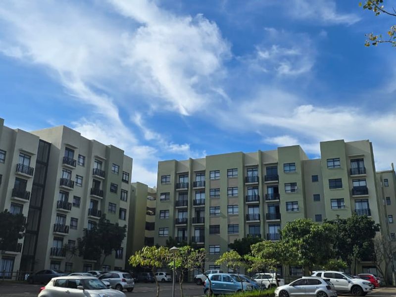 Exterior view of a multi-story apartment complex featuring balconies and an adjacent paved parking area under a clear blue sky.