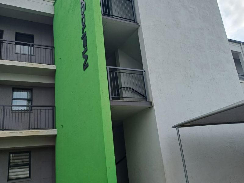 Exterior view of a multi-story apartment building featuring a green accent facade, metal balcony railings, and a paved entrance walkway.
