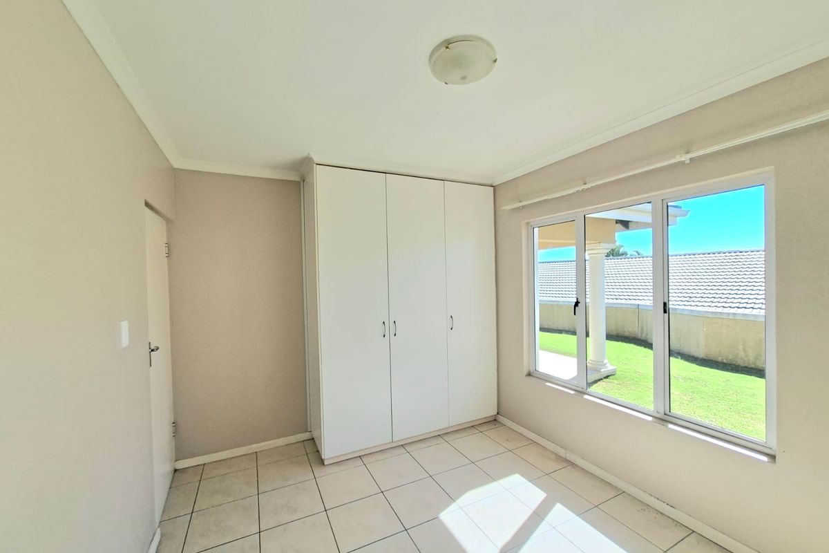 A bright, empty bedroom featuring neutral tiled flooring, built-in white cupboards, and large windows providing natural light and garden views.