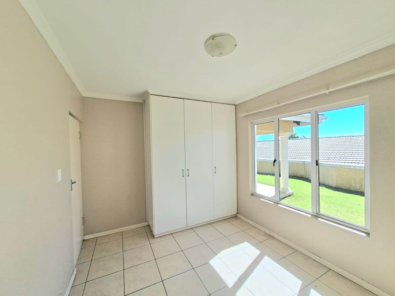 A bright, empty bedroom featuring neutral tiled flooring, built-in white cupboards, and large windows providing natural light and garden views.