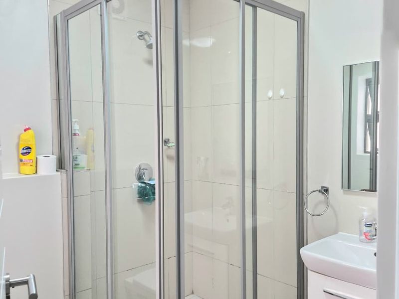 A functional bathroom featuring a corner shower enclosure, wall-mounted basin with vanity storage, and neutral wall tiling.