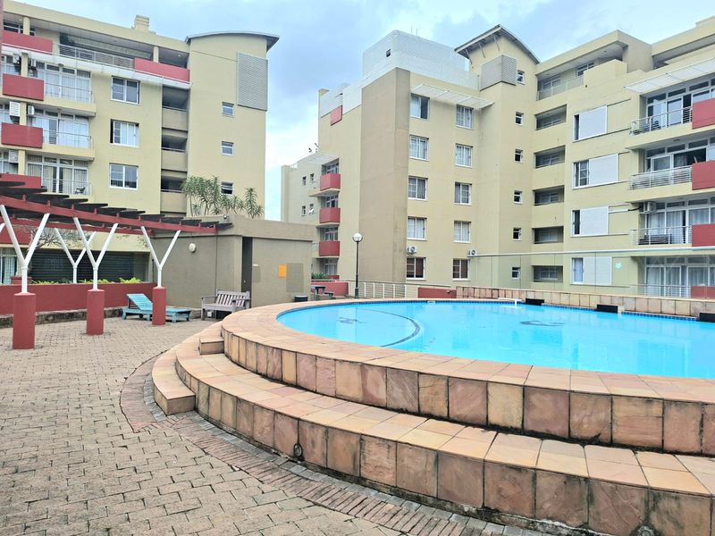 Communal outdoor pool area featuring a circular swimming pool with stone coping, paved terrace, and a pergola structure within an apartment complex.