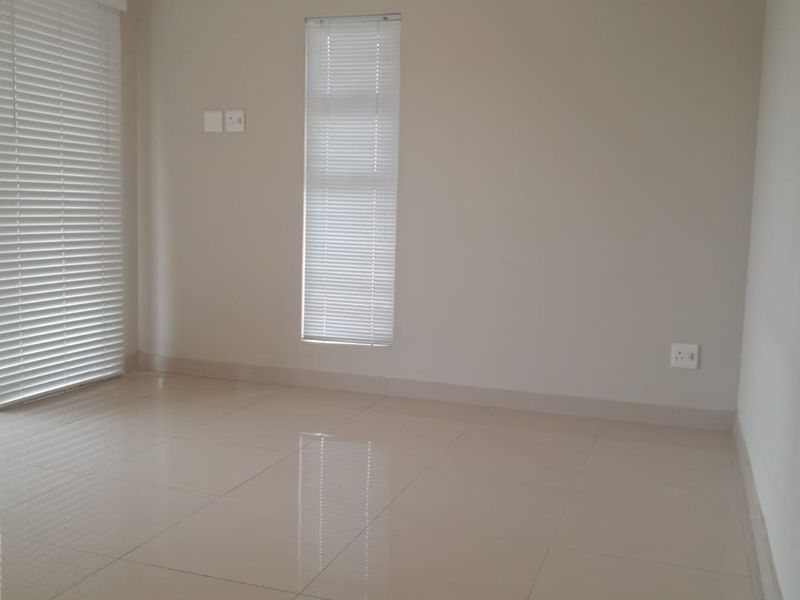 A bright, empty living area featuring neutral tiled flooring, clean walls, and fitted venetian blinds, offering a versatile space ready for furnishing.