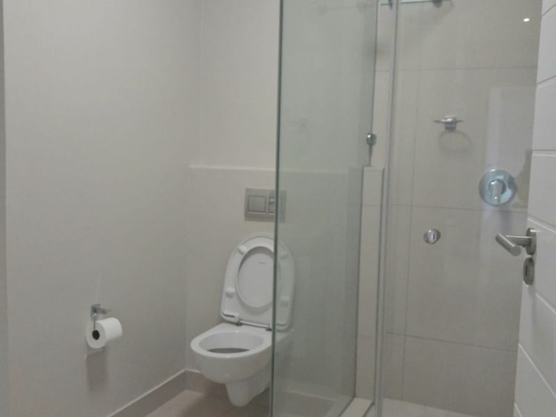 A clean, modern bathroom featuring a wall-mounted toilet, a glass-enclosed shower, and neutral floor tiling.