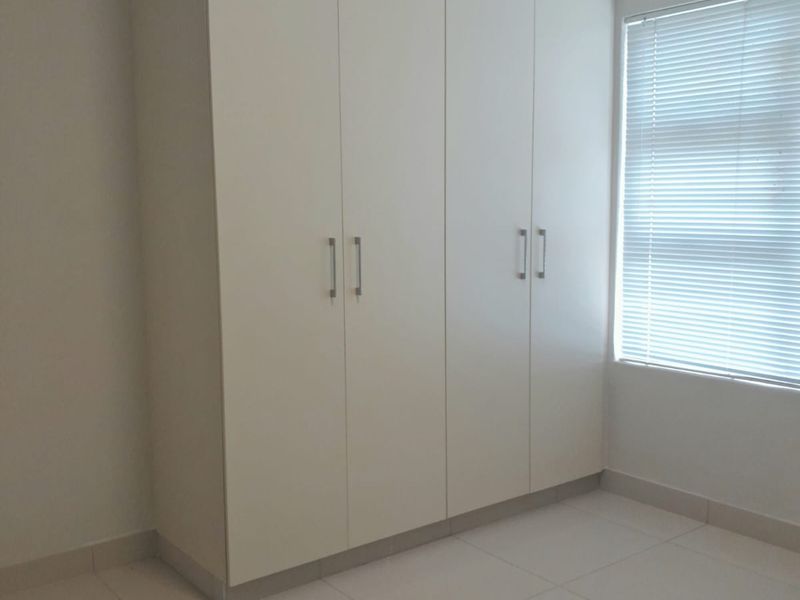 A clean, empty bedroom featuring neutral tiled flooring, built-in floor-to-ceiling cupboards, and venetian blinds on the window.