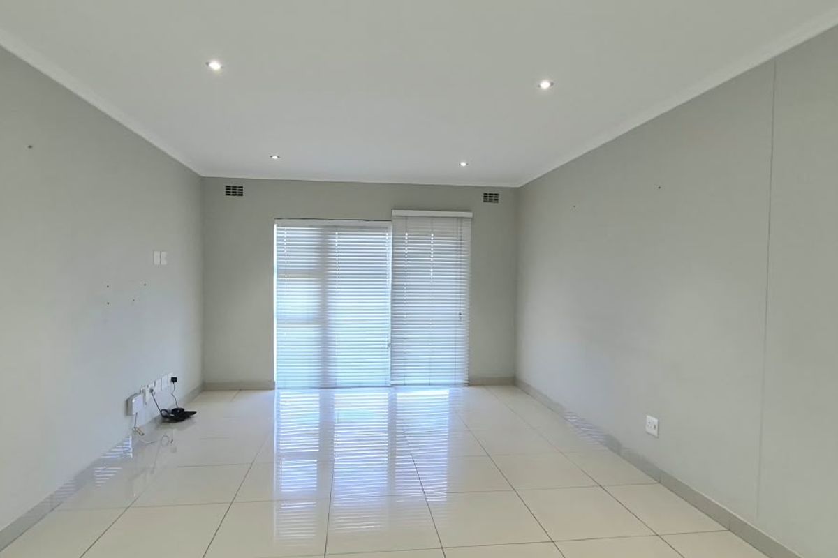 A bright, empty living room featuring neutral tiled flooring, recessed downlighting, and a sliding glass door with fitted venetian blinds.