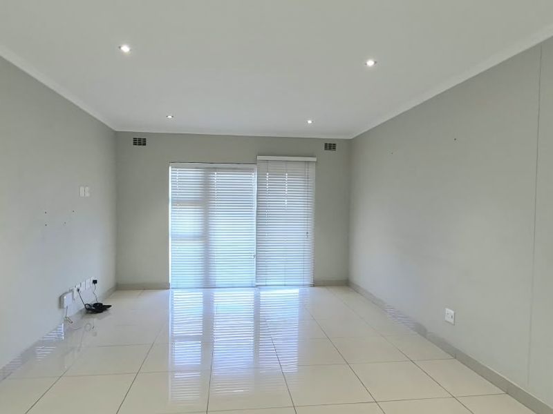 A bright, empty living room featuring neutral tiled flooring, recessed downlighting, and a sliding glass door with fitted venetian blinds.
