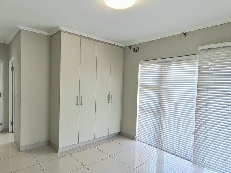 A bright, empty bedroom featuring neutral tiled flooring, built-in cupboards, and large windows fitted with venetian blinds.