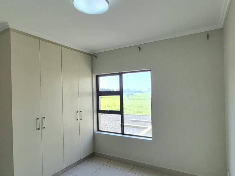 A clean, empty bedroom featuring neutral tiled flooring, built-in cupboards, and an aluminium window with a view of the surrounding landscape.