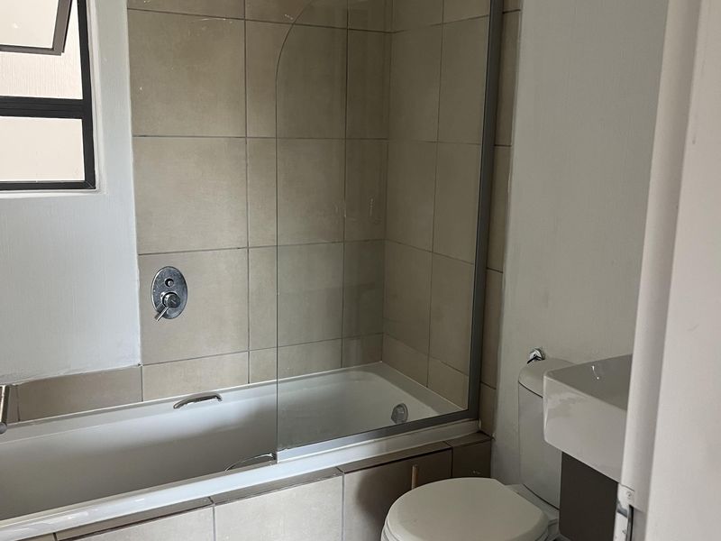 A functional bathroom featuring a bathtub with a glass shower screen, a wall-mounted basin, and neutral floor and wall tiling.