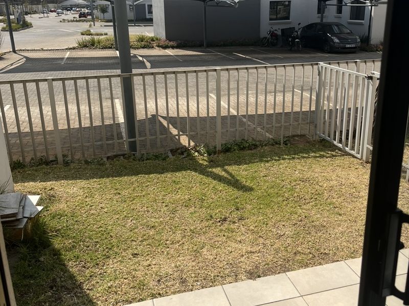 A private ground floor patio area featuring a small lawn, tiled flooring, and a metal fence, overlooking a communal parking area with shade cloth carports.