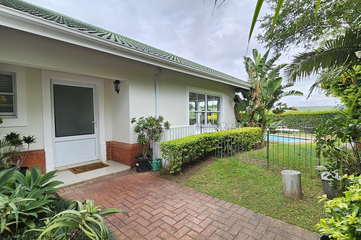 A well-maintained rear exterior view of the townhouse featuring a brick-paved patio, landscaped garden, and a glimpse of the swimming pool.