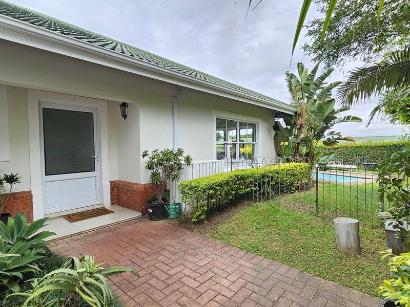 A well-maintained rear exterior view of the townhouse featuring a brick-paved patio, landscaped garden, and a glimpse of the swimming pool.