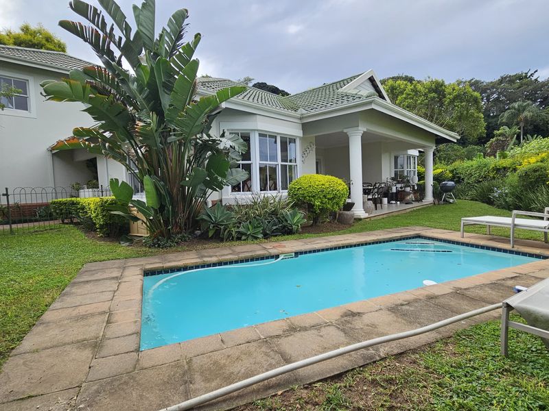 A rear exterior view of the townhouse featuring a swimming pool, paved surround, and a covered patio area set within a landscaped garden.