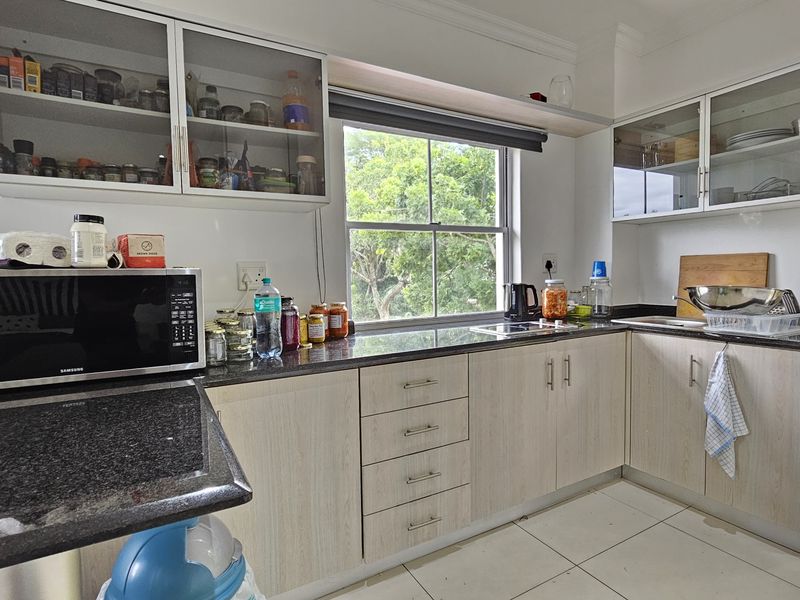 A functional kitchen featuring granite countertops, light wood-grain cabinetry, glass-front upper storage, and tiled flooring with a window overlooking greenery.