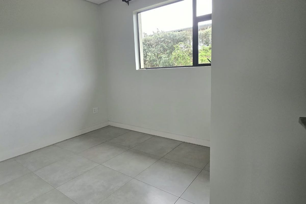 A clean, empty bedroom featuring neutral grey floor tiles, white walls, and a large window providing natural light and a garden view.
