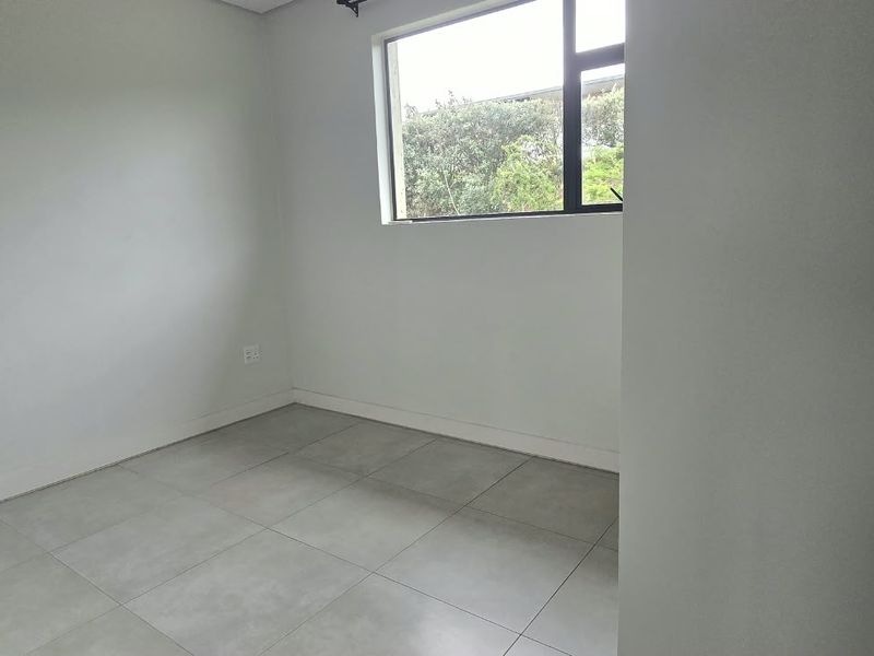 A clean, empty bedroom featuring neutral grey floor tiles, white walls, and a large window providing natural light and a garden view.