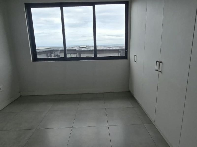 A bright, empty bedroom featuring neutral tiled flooring, ample built-in cupboard storage, and a large window offering natural light.