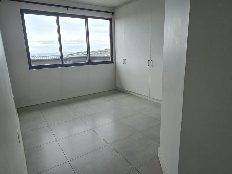 A bright, empty bedroom featuring neutral tiled flooring, built-in cupboards, and large windows offering natural light and exterior views.