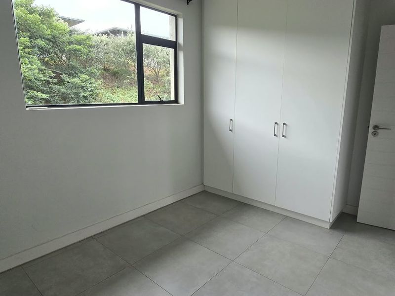 A bright, empty bedroom featuring modern grey floor tiles, built-in white cupboards, and a window overlooking greenery.