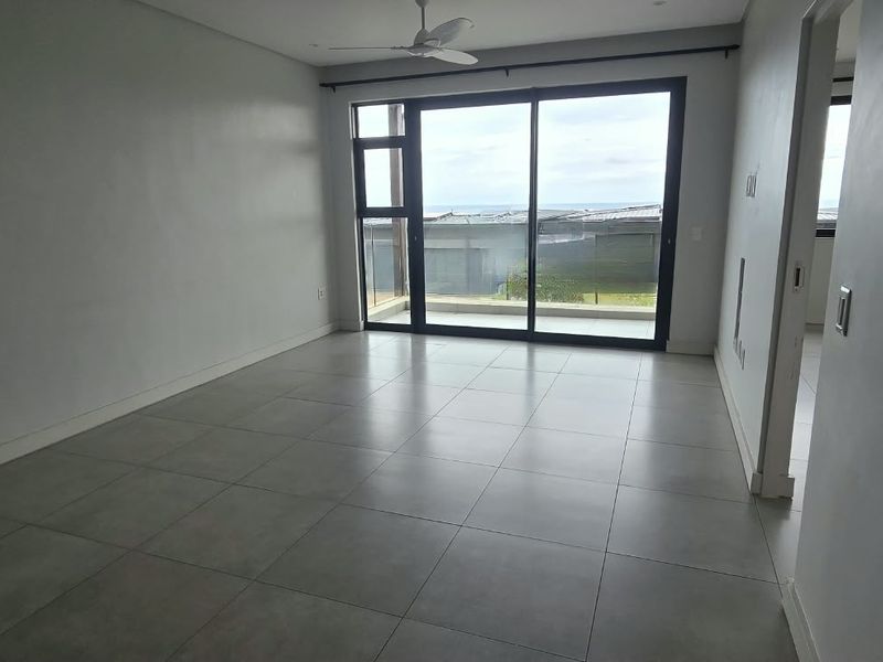 An empty, well-lit living room featuring large sliding glass doors, neutral tiled flooring, and a ceiling fan, offering a clean and modern aesthetic.