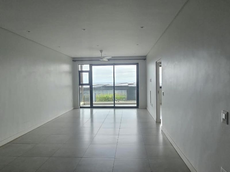 A spacious, empty living area featuring neutral tiled flooring, recessed lighting, and a large sliding glass door providing natural light and outdoor access.