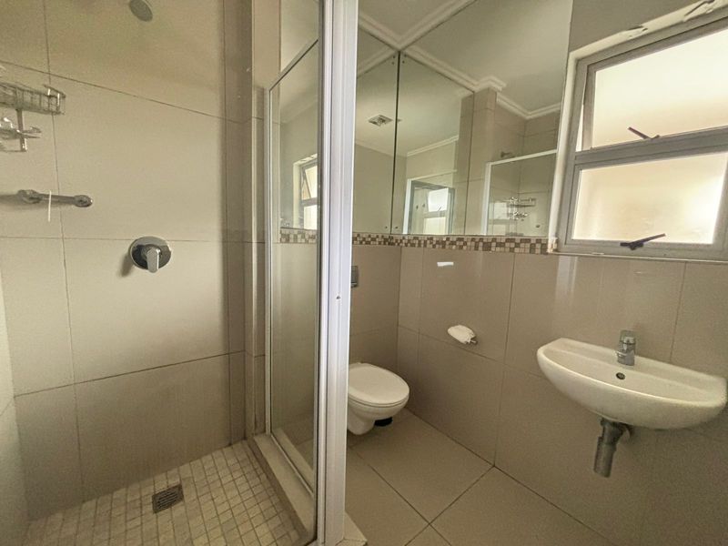 A functional bathroom featuring a glass-enclosed shower, wall-mounted basin, toilet, and large mirror cabinet with neutral tiled finishes.