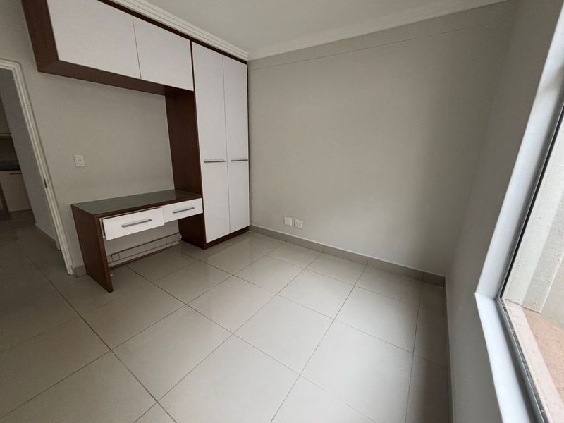 A bright, empty bedroom featuring neutral tiled flooring, a built-in wardrobe, and an integrated desk unit with overhead storage cabinetry.