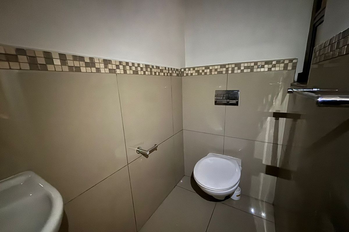 A clean, modern guest bathroom featuring a wall-mounted toilet, neutral tiled walls with a decorative mosaic border, and a functional wash basin.