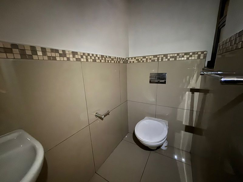 A clean, modern guest bathroom featuring a wall-mounted toilet, neutral tiled walls with a decorative mosaic border, and a functional wash basin.