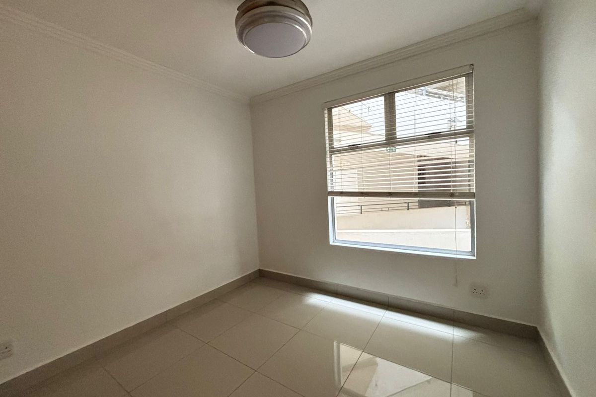A clean, empty bedroom featuring neutral tiled flooring, a ceiling fan, and a window fitted with venetian blinds.