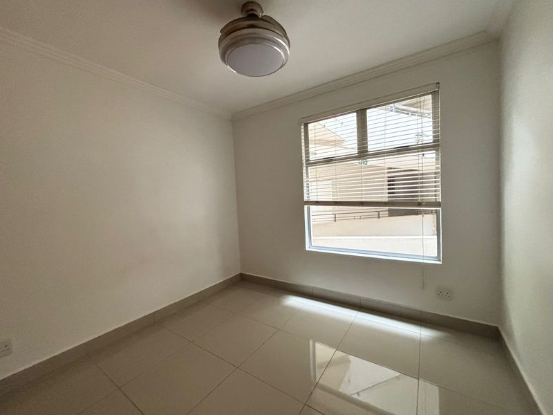 A clean, empty bedroom featuring neutral tiled flooring, a ceiling fan, and a window fitted with venetian blinds.
