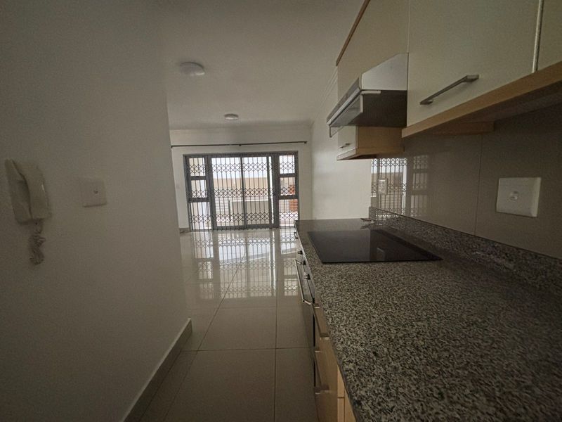 A clean, modern kitchen featuring granite countertops, built-in cabinetry, and an electric hob, leading into an open-plan living area with security gate access.