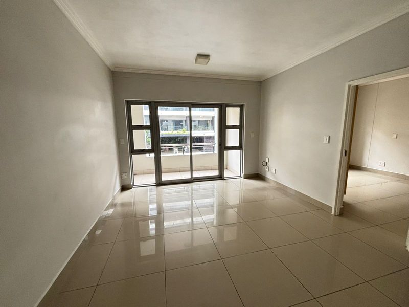 A bright, empty living room featuring neutral tiled flooring, a sliding glass door leading to a balcony, and clean, well-maintained wall finishes.