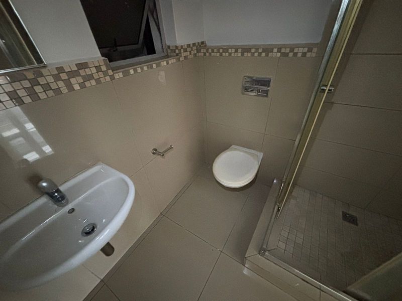 A functional bathroom featuring a wall-mounted basin, wall-hung toilet, and a glass-enclosed shower with neutral tiled finishes and a decorative mosaic border.