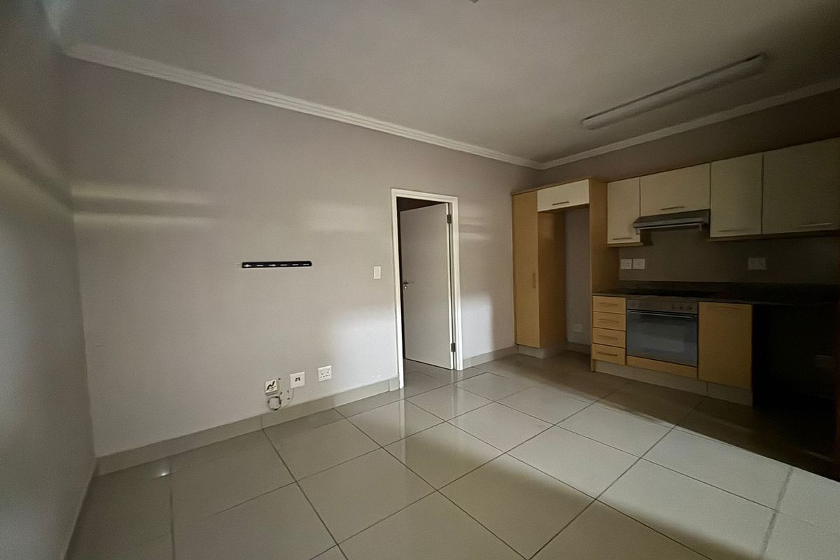 An open-plan living and kitchen area featuring tiled flooring, built-in cabinetry, and integrated kitchen appliances in a neutral, vacant space.