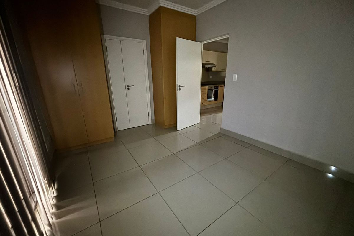 A clean, empty bedroom featuring neutral tiled flooring, built-in wooden cupboards, and direct access to the kitchen area.