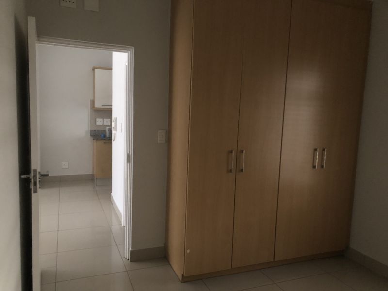 A bedroom featuring built-in wooden cupboards and tiled flooring, with a doorway leading to an adjacent kitchen area.