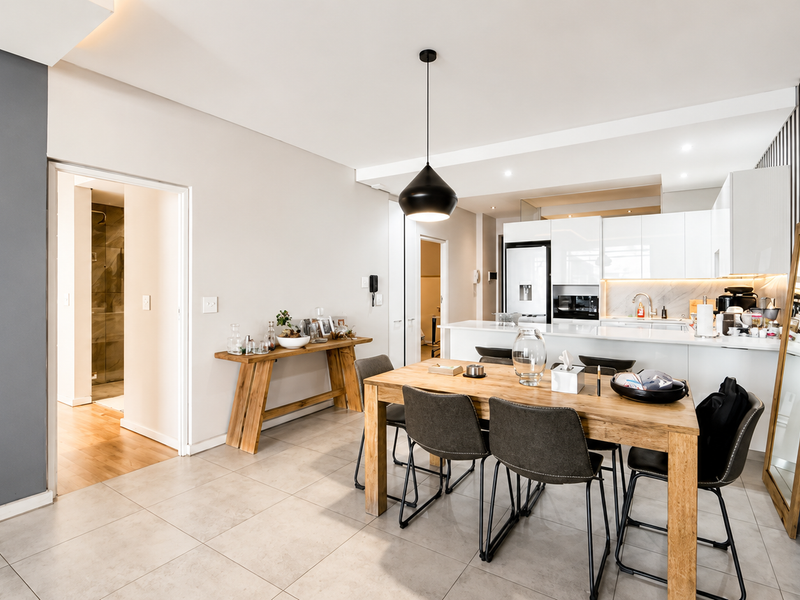 Modern open-plan living and dining area featuring a sleek kitchen with white cabinetry, stone countertops, and contemporary lighting, seamlessly connected to the dining space.