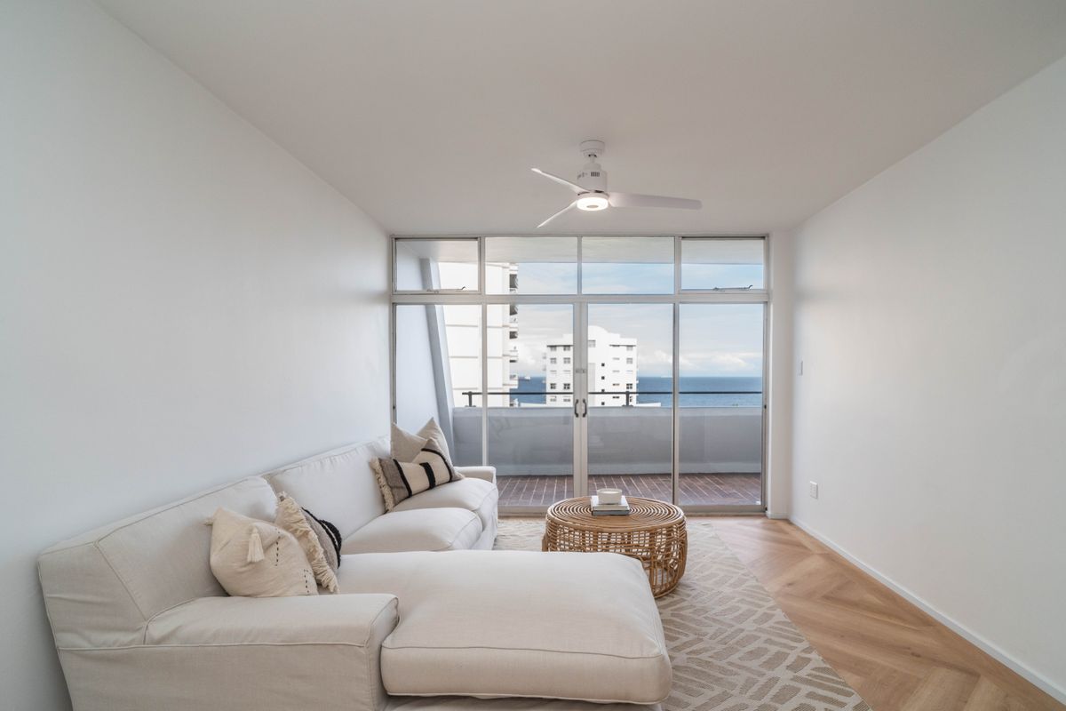 Bright living room featuring herringbone timber flooring, a modern ceiling fan, and large sliding glass doors opening onto a balcony with ocean views.