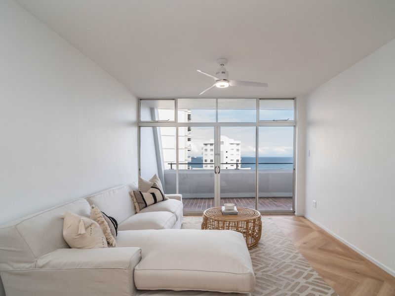 Bright living room featuring herringbone timber flooring, a modern ceiling fan, and large sliding glass doors opening onto a balcony with ocean views.