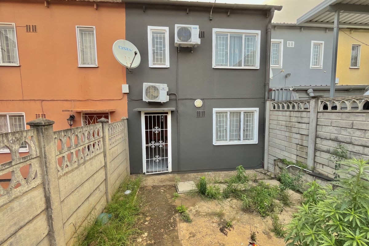 Rear exterior view of a townhouse featuring a small garden area, security gate, and wall-mounted air conditioning units.