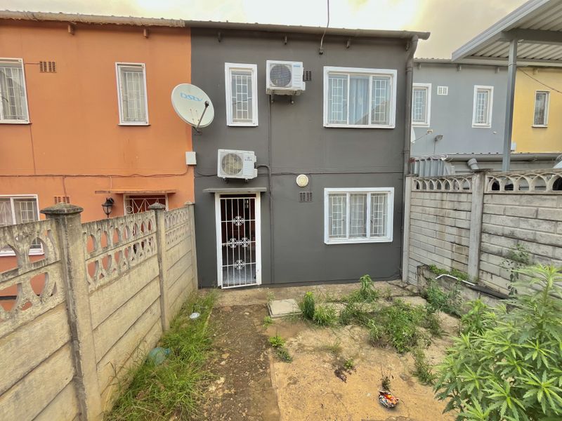 Rear exterior view of a townhouse featuring a small garden area, security gate, and wall-mounted air conditioning units.