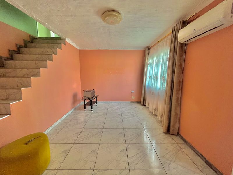 A spacious living area featuring tiled flooring, a built-in staircase, and a wall-mounted air conditioning unit, offering a blank canvas for interior design.